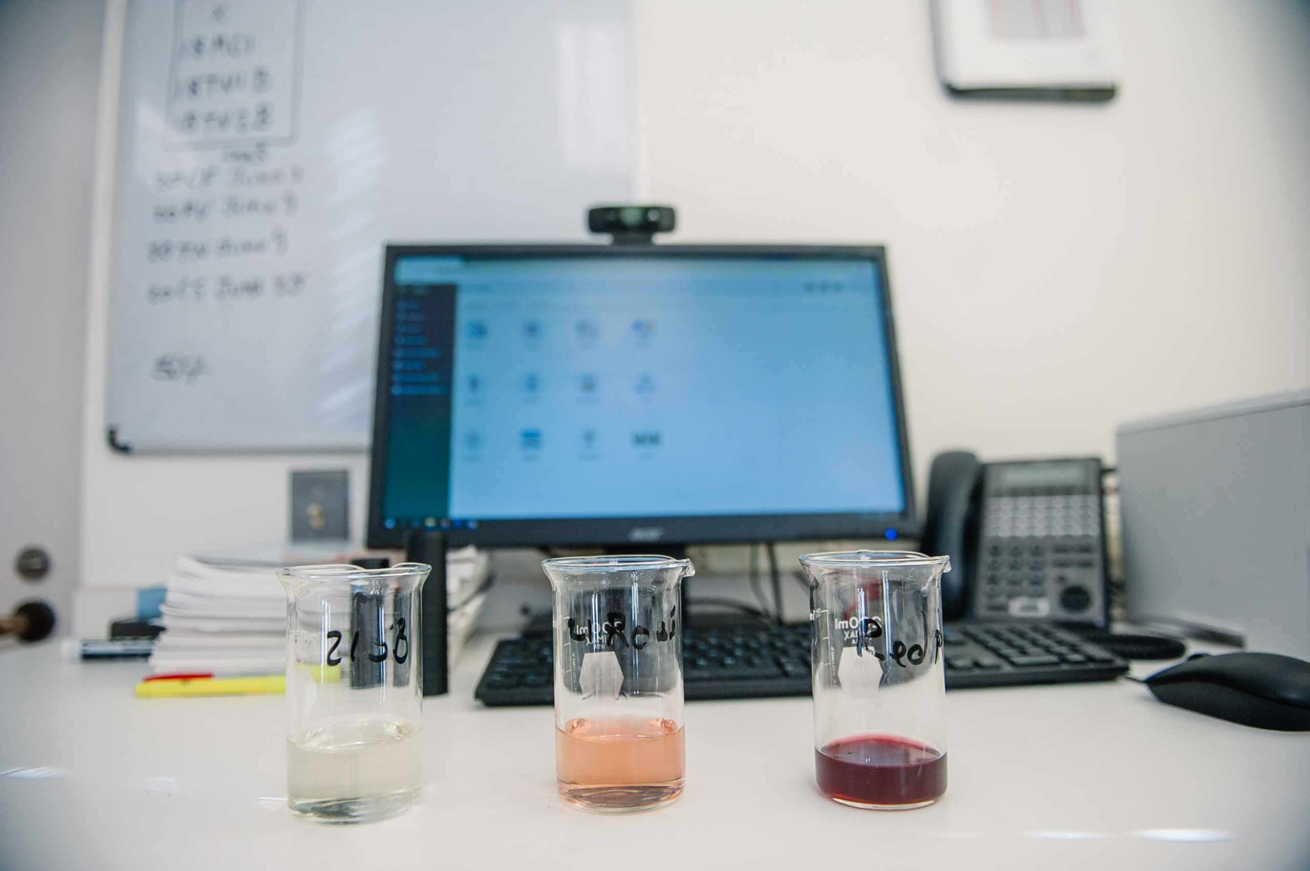 Three glass beakers filled with white, red, and pink wines on a desk in front of a computer screen of winery software