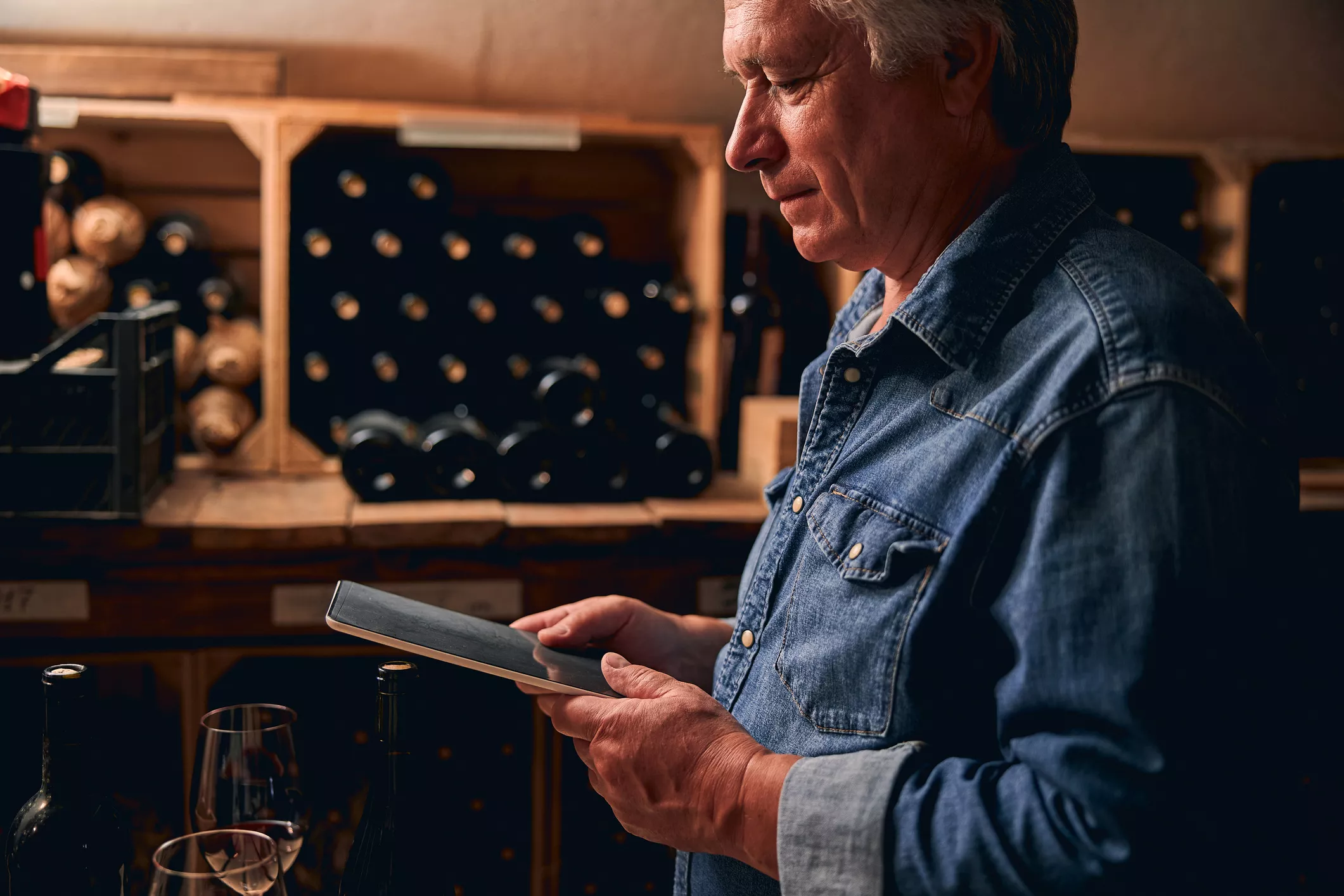 Wine cellar worker using wholesale distribution software to manage winery sales orders, surrounded by bottled wine